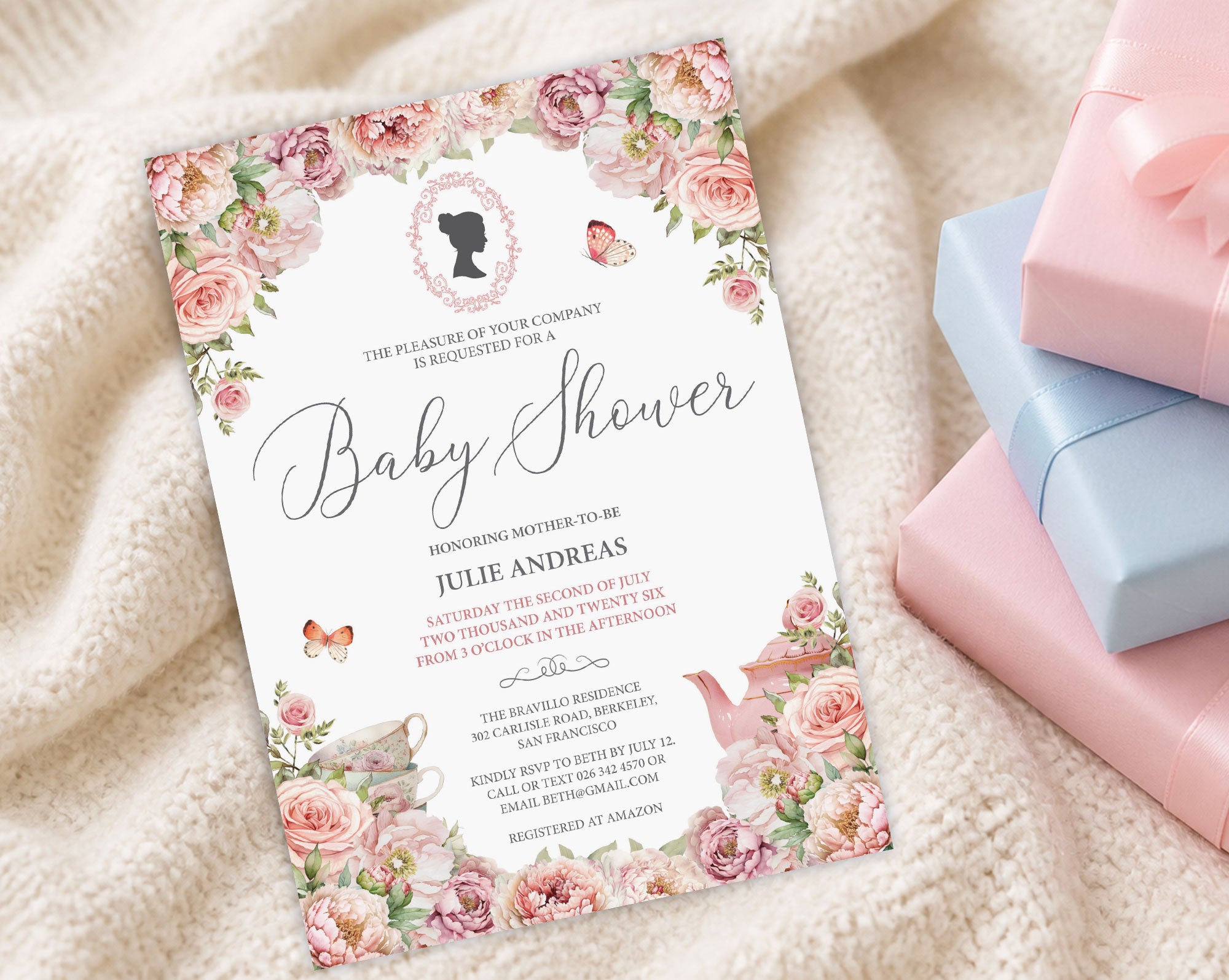 a blush pink baby shower invitation with cameo and flowers on a cream baby blanket next to some pink and blue wrapped gifts