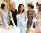 5 ladies at a baby showing wearing blue pastel dresses and white gloves with a pregnant lady in the middle hold a pair of white baby booties, next to a tea table with food.