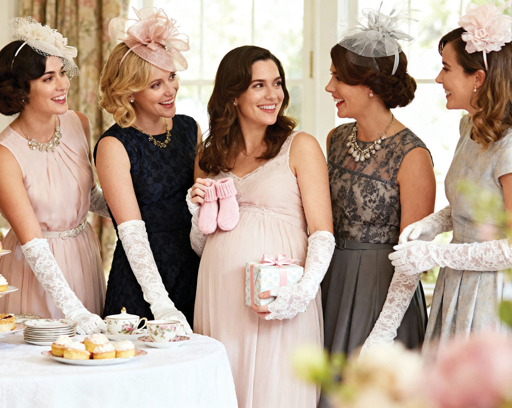 Five women at a pastel baby shower in elegant dresses with hats and gloves, standing around a table with tea and pastries.