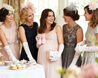 Five women at a pastel baby shower in elegant dresses with hats and gloves, standing around a table with tea and pastries.