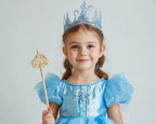A young girl with braided hair wearing a light blue princess costume, matching tiara, and holding a gold scepter against a plain light gray background.