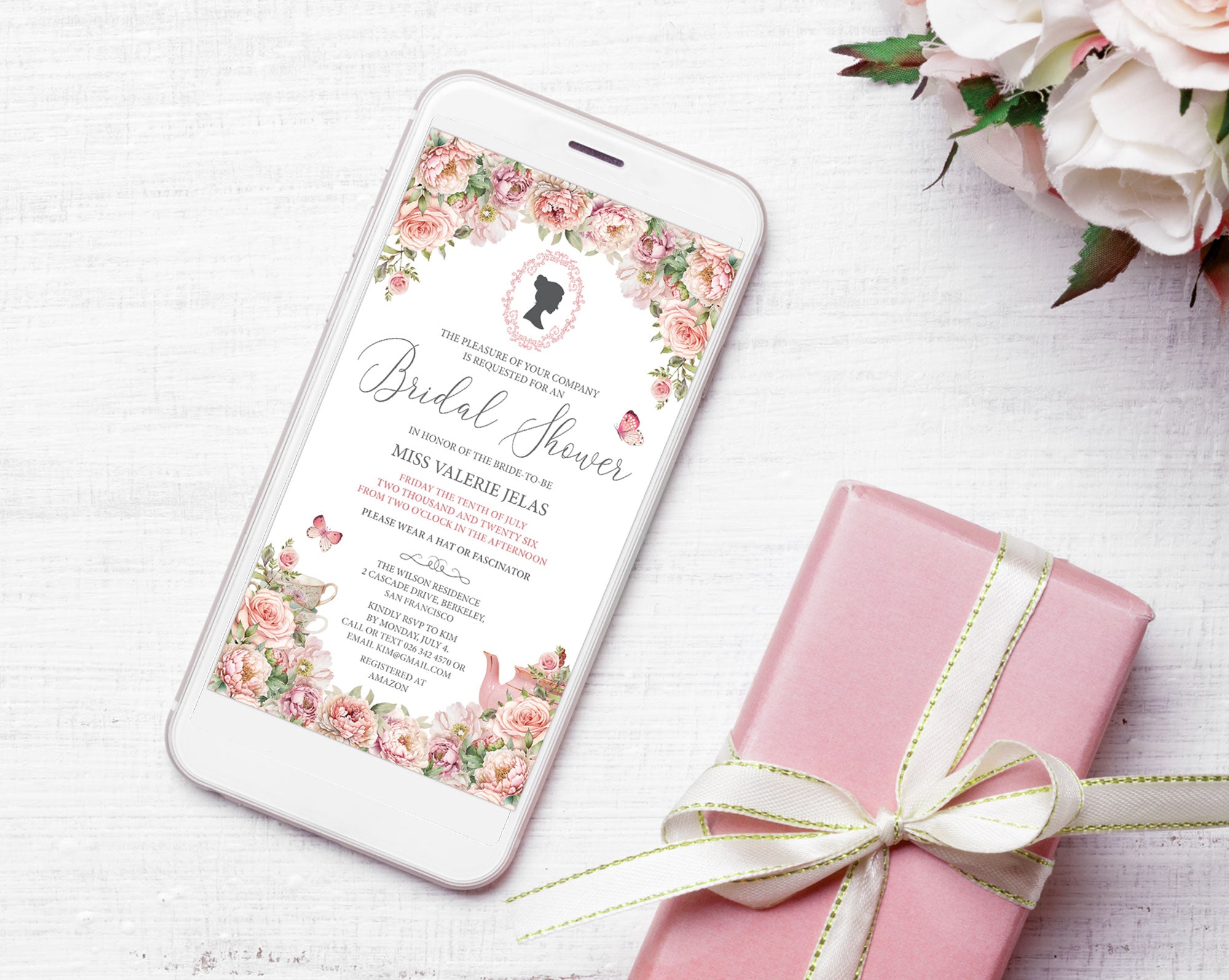 Pretty blush pink flower Bridal Shower Mobile invitation with Bridgerton Style cameo and butterflies showing on the screen of a white mobile phone, laying on a white background next to some white roses and a pink wrapped gift