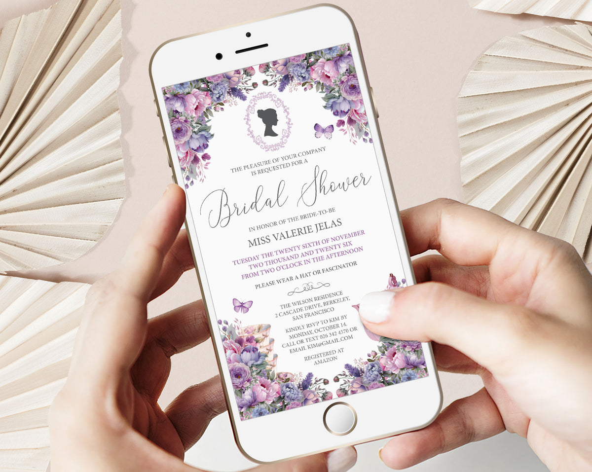 A person holds a white smartphone displaying a bridal shower invitation with purple floral borders and elegant script text. The scene is styled with beige paper fans on a soft pink background.