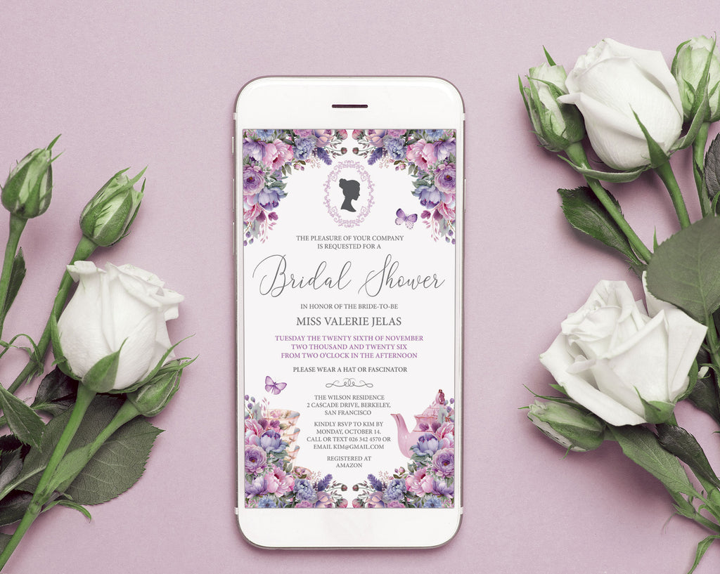 A smartphone lying on a dusty pink surface, displaying a bridal shower invitation with purple and lavender floral borders and elegant script text. White roses and rose buds are arranged around the phone in a styled flat-lay composition.