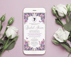 A smartphone lying on a dusty pink surface, displaying a bridal shower invitation with purple and lavender floral borders and elegant script text. White roses and rose buds are arranged around the phone in a styled flat-lay composition.