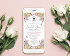 A smartphone lying on a soft pink surface, displaying a pastel flower bridal shower invitation design. White roses and rose buds are arranged around the phone, creating a styled flat-lay composition.