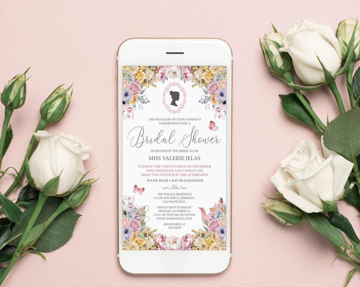 A smartphone lying on a soft pink surface, displaying a pastel flower bridal shower invitation design. White roses and rose buds are arranged around the phone, creating a styled flat-lay composition.