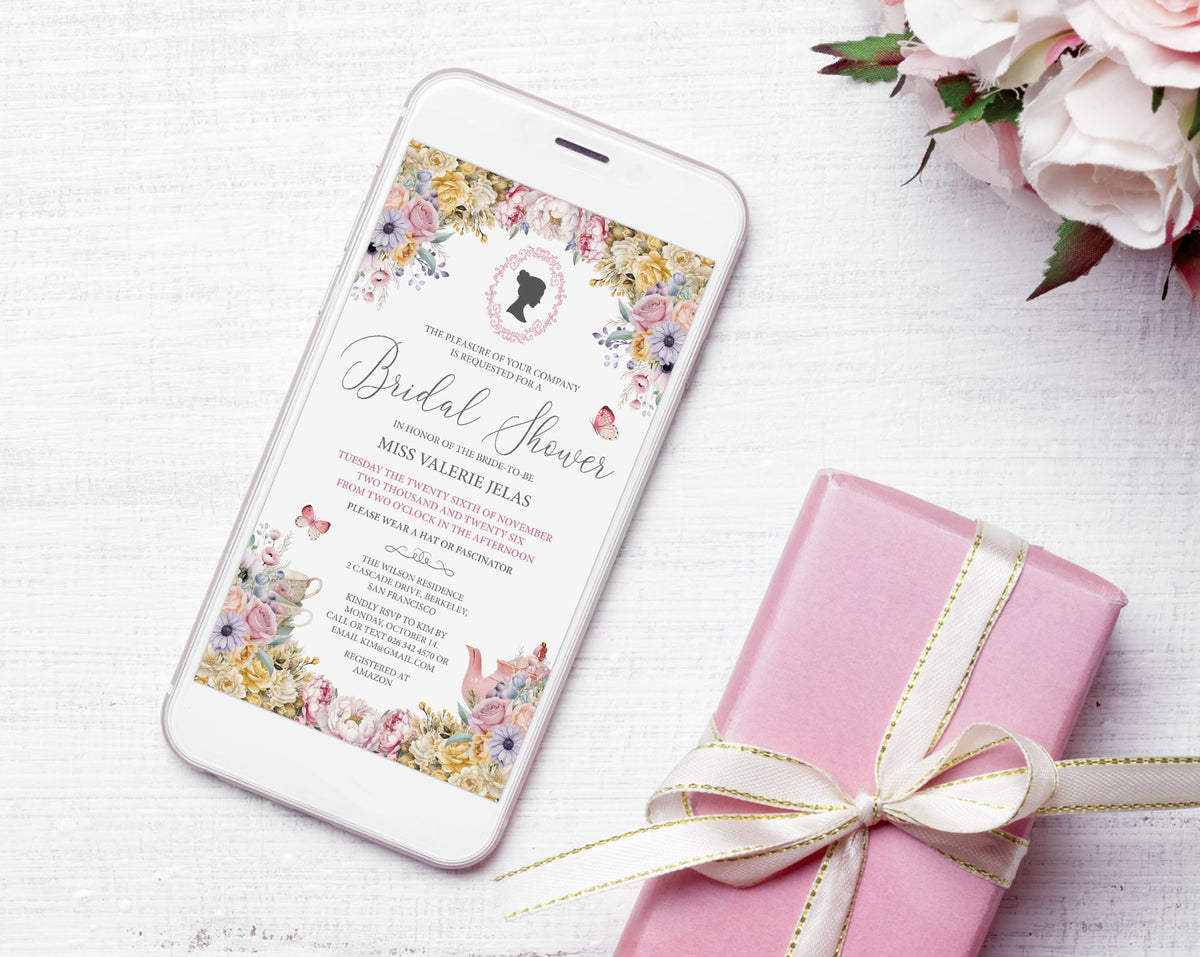 A smartphone displaying a floral bridal shower invitation, styled on a white wood surface next to a pink gift box tied with a cream ribbon and pink flowers in the corner.