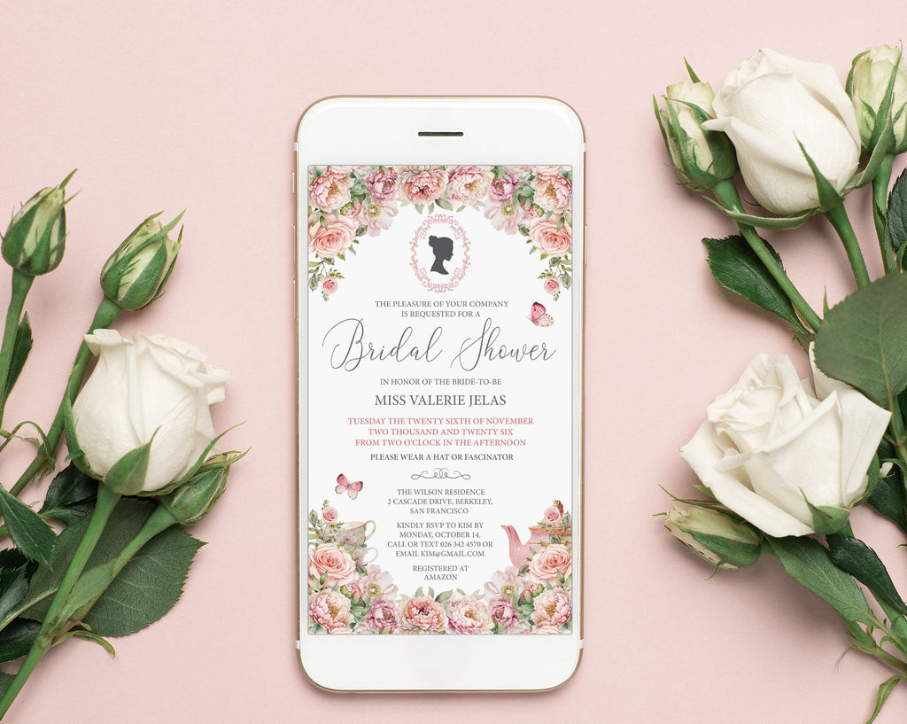A smartphone lying on a soft pink surface, displaying a regency style blush pink floral bridal shower invitation design. White roses and rose buds are arranged around the phone, creating a styled flat-lay composition.
