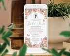 A mobile phone showing a Regency Bridal Tea blush pink Invitation with teapot and teacups, cameo and butterflies, leaning up against a vase with greenery