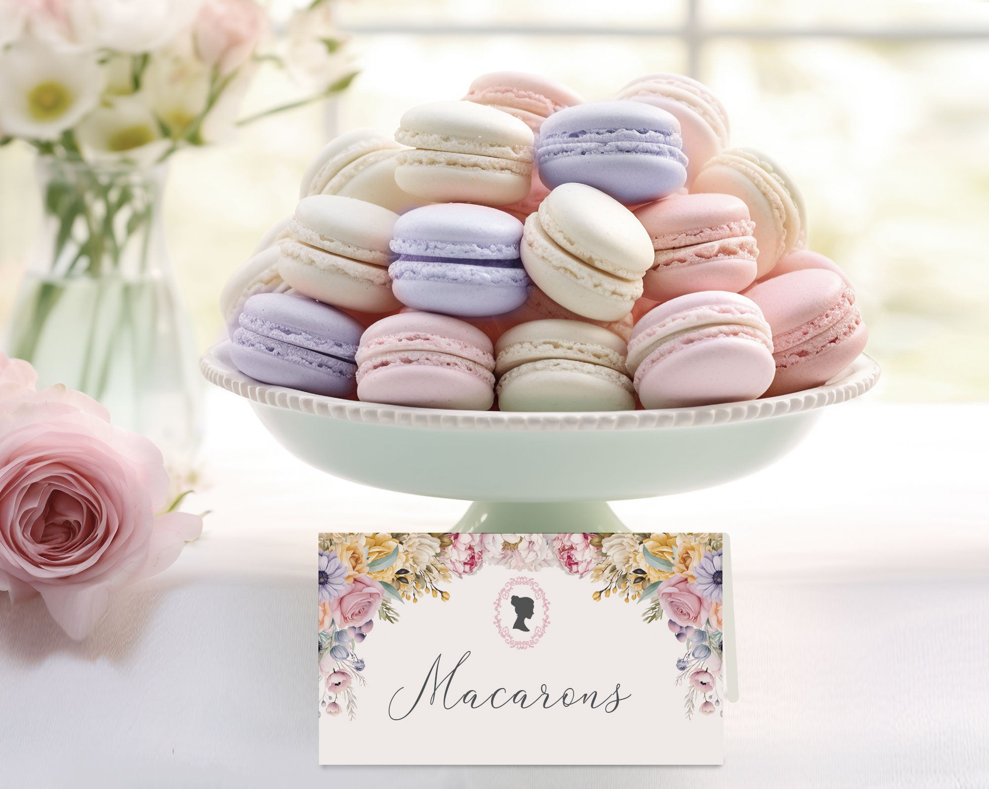 Pastel macarons on a white stand with a pastel floral name card below, set against a blurred floral background.
