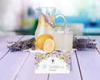 Lavender lemonade with a glass and pitcher on a wooden surface, featuring a drink name tent card in the Bridgerton style