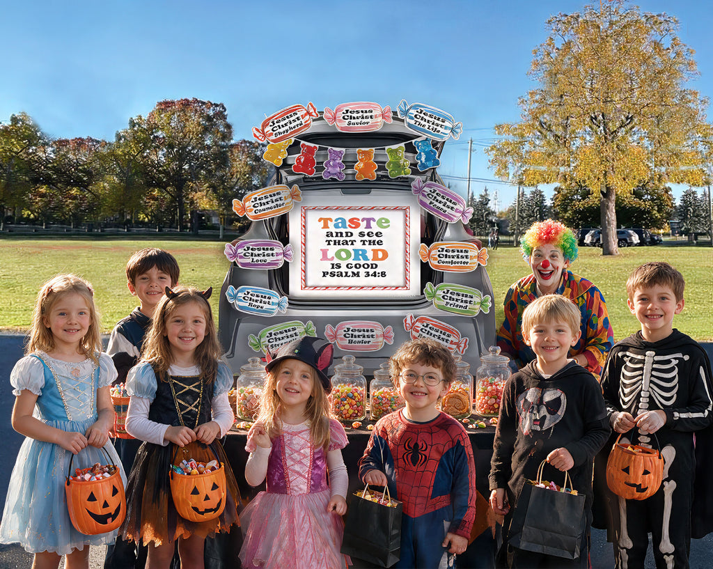 A group of children in Halloween costumes stand in front of a large candy cart outdoors on a grassy park area under a blue sky. One child is dressed as a clown.