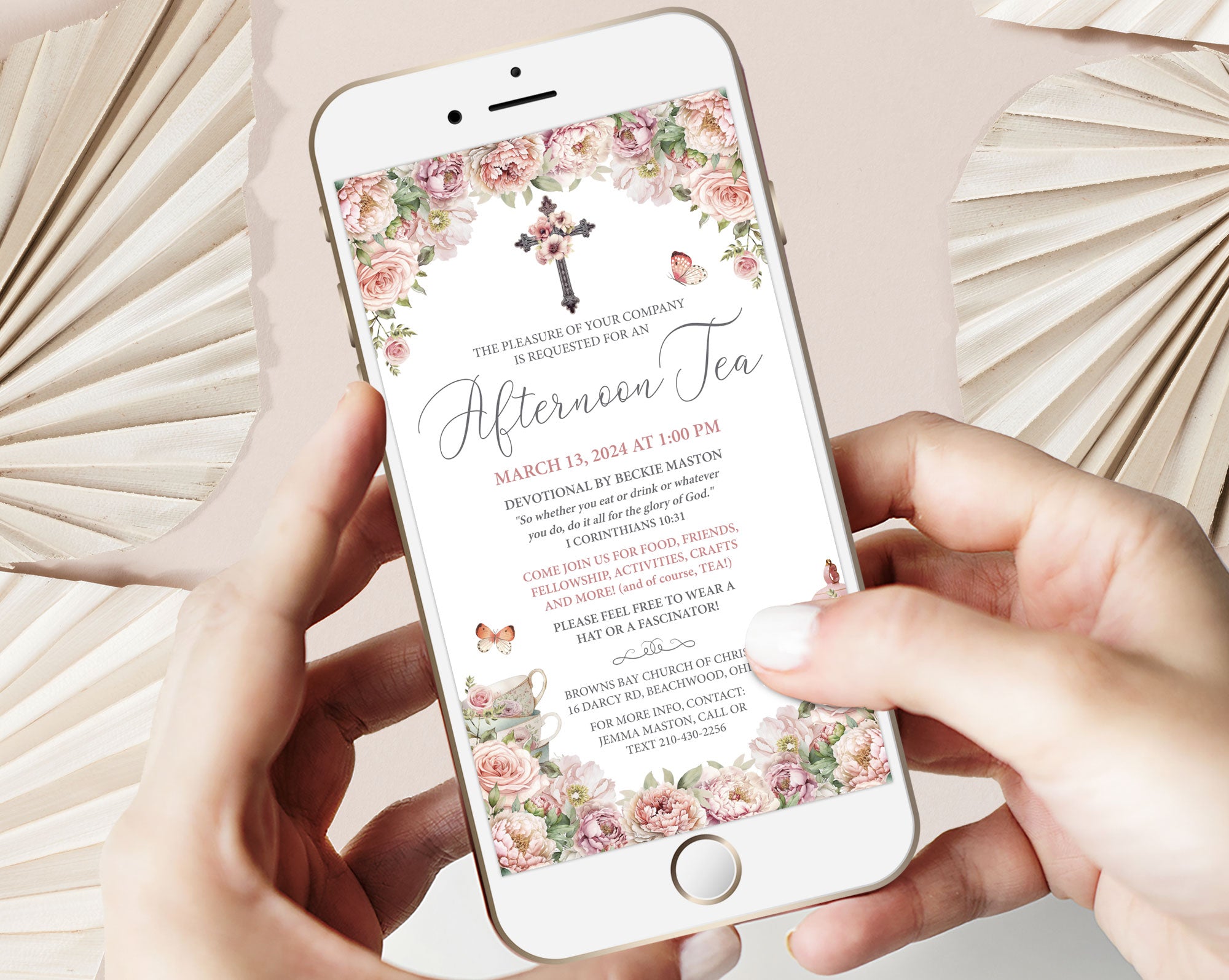 Phone displaying a blush pink flower afternoon tea invitation with floral cross held by hands on a light background