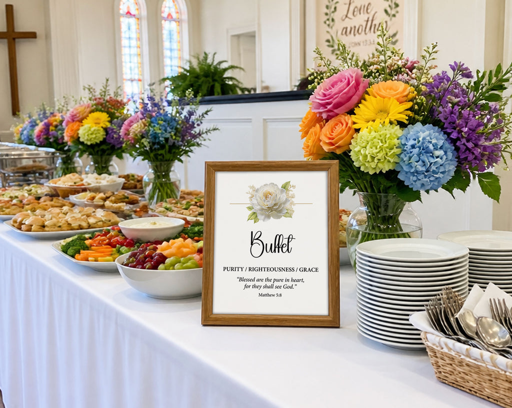 a buffet sign in a wooden frame with a white flower and scripture, on a long white table with food, rainbow flowers, stacked white plates and cutlery