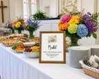 a buffet sign in a wooden frame with a white flower and scripture, on a long white table with food, rainbow flowers, stacked white plates and cutlery