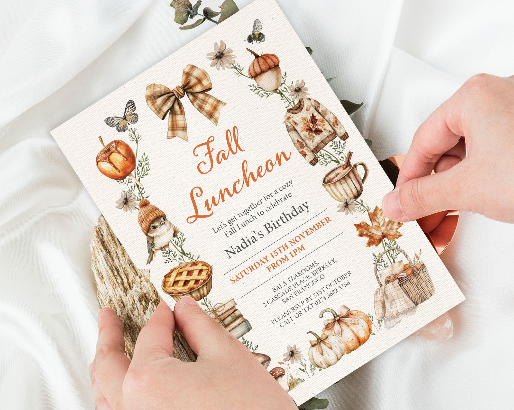 A person holds a printed fall-themed birthday invitation card with autumn illustrations, including pumpkins, acorns, leaves, and bows, over a soft white fabric background.