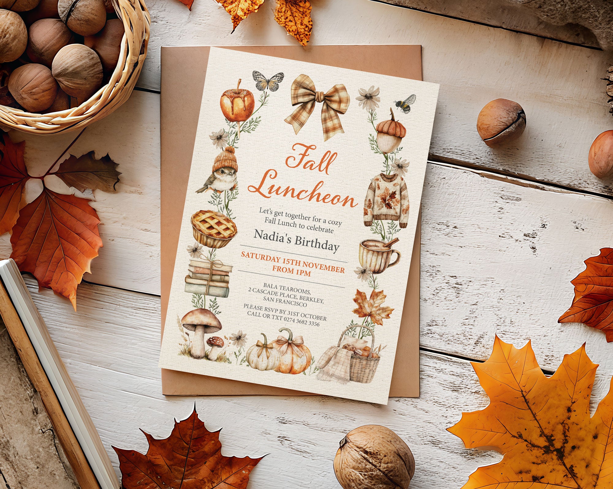 A fall-themed birthday invitation card with autumn illustrations, placed on a kraft envelope over a rustic white wood surface. The scene is styled with orange leaves, acorns, and a basket of walnuts.