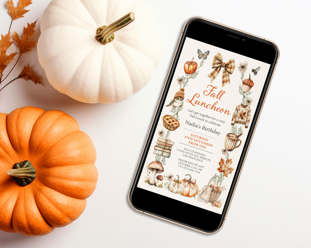 A smartphone lying on a white surface displays a fall-themed birthday invitation design with illustrations of pumpkins, bows, leaves, and autumn motifs. The phone is styled with real white and orange pumpkins and dried leaves around it.