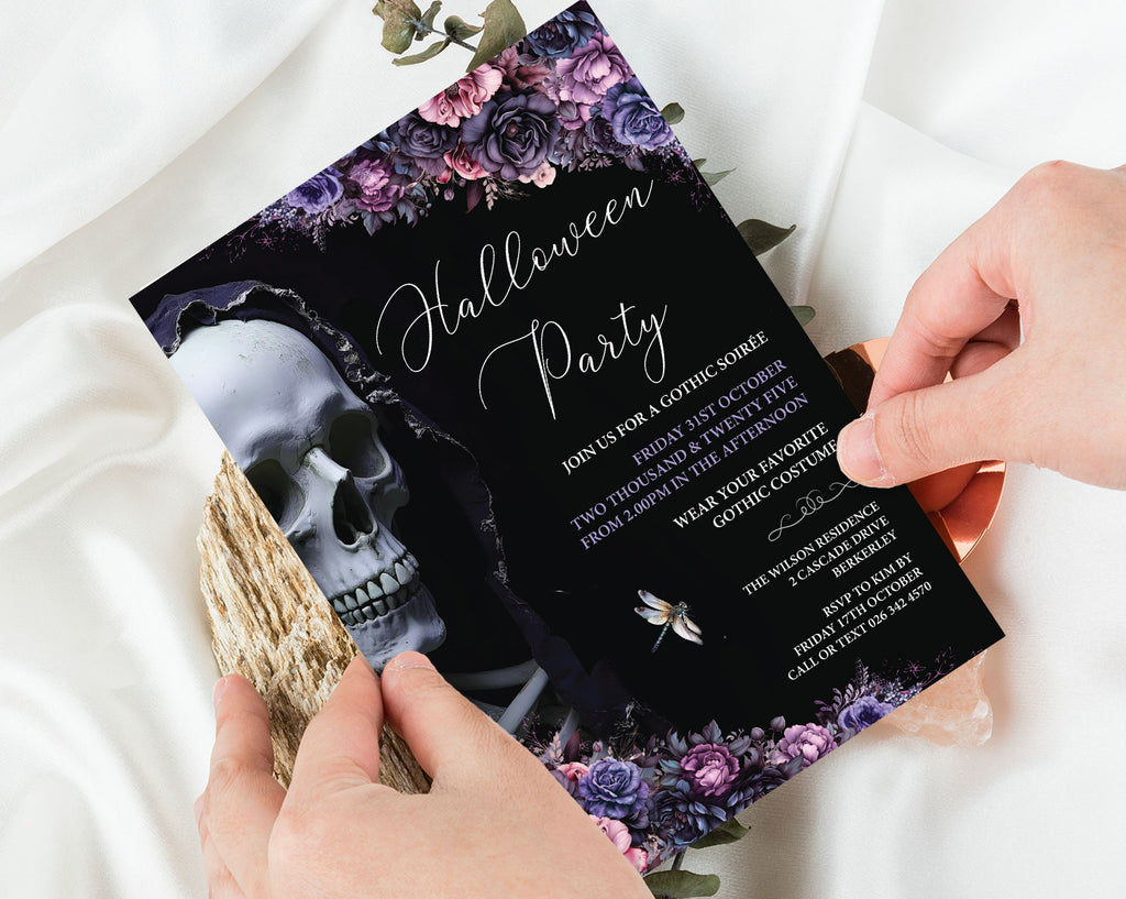 A gothic Halloween party invitation card being held by two hands over a white satin fabric background. The invitation features a black background with a skull illustration, purple and pink floral borders, and elegant white script text reading 'Halloween Party'.