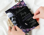 A gothic Halloween party invitation card being held by two hands over a white satin fabric background. The invitation features a black background with a skull illustration, purple and pink floral borders, and elegant white script text reading 'Halloween Party'.