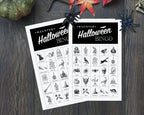 Two printable Halloween bingo cards laid on a dark wood surface, with decorative spiderwebs, a pumpkin, and autumn leaves around them. The cards are white with black headers reading 'Haunted Halloween Bingo' and a grid of black Halloween-themed icons and words.