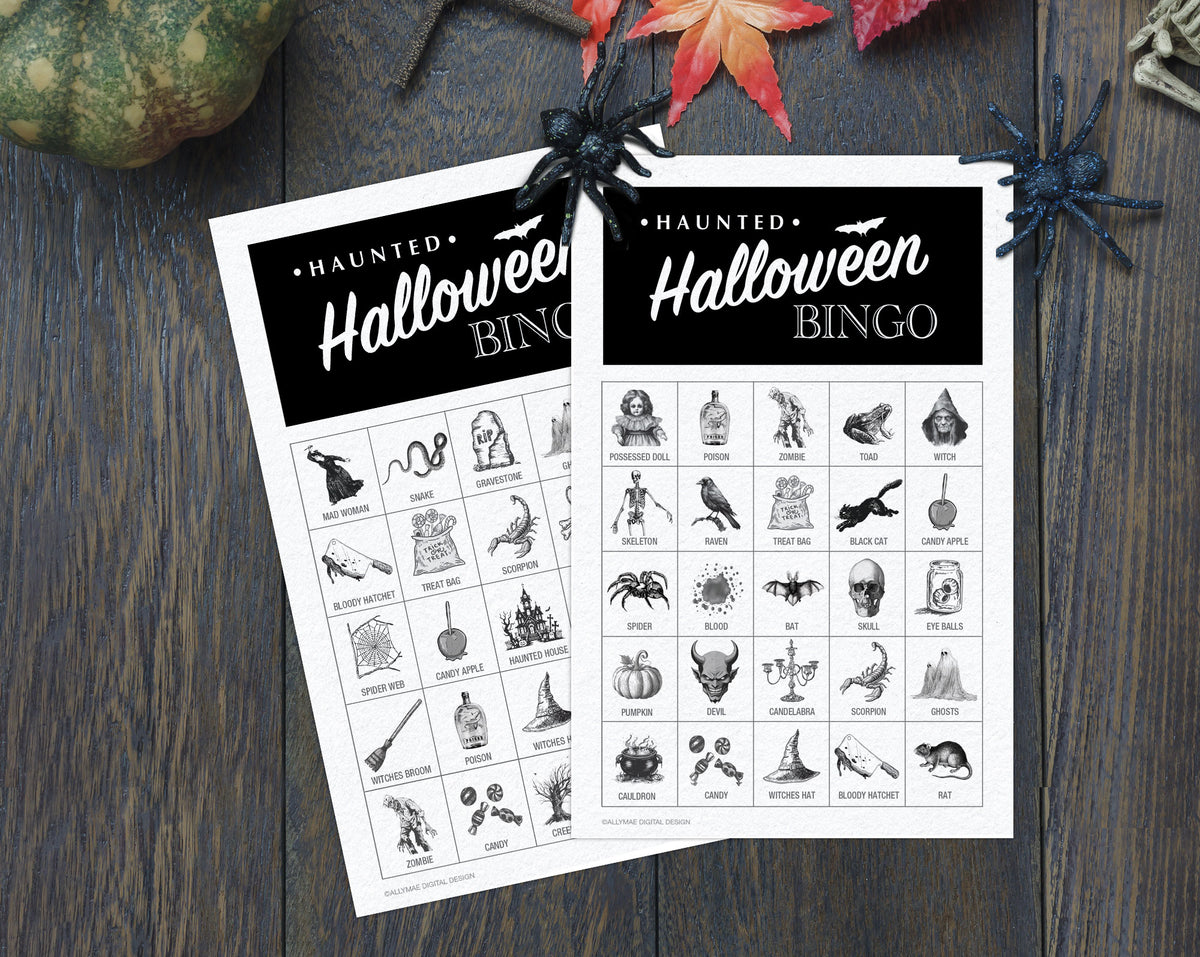 Two printable Halloween bingo cards laid on a dark wood surface, with decorative spiderwebs, a pumpkin, and autumn leaves around them. The cards are white with black headers reading 'Haunted Halloween Bingo' and a grid of black Halloween-themed icons and words.