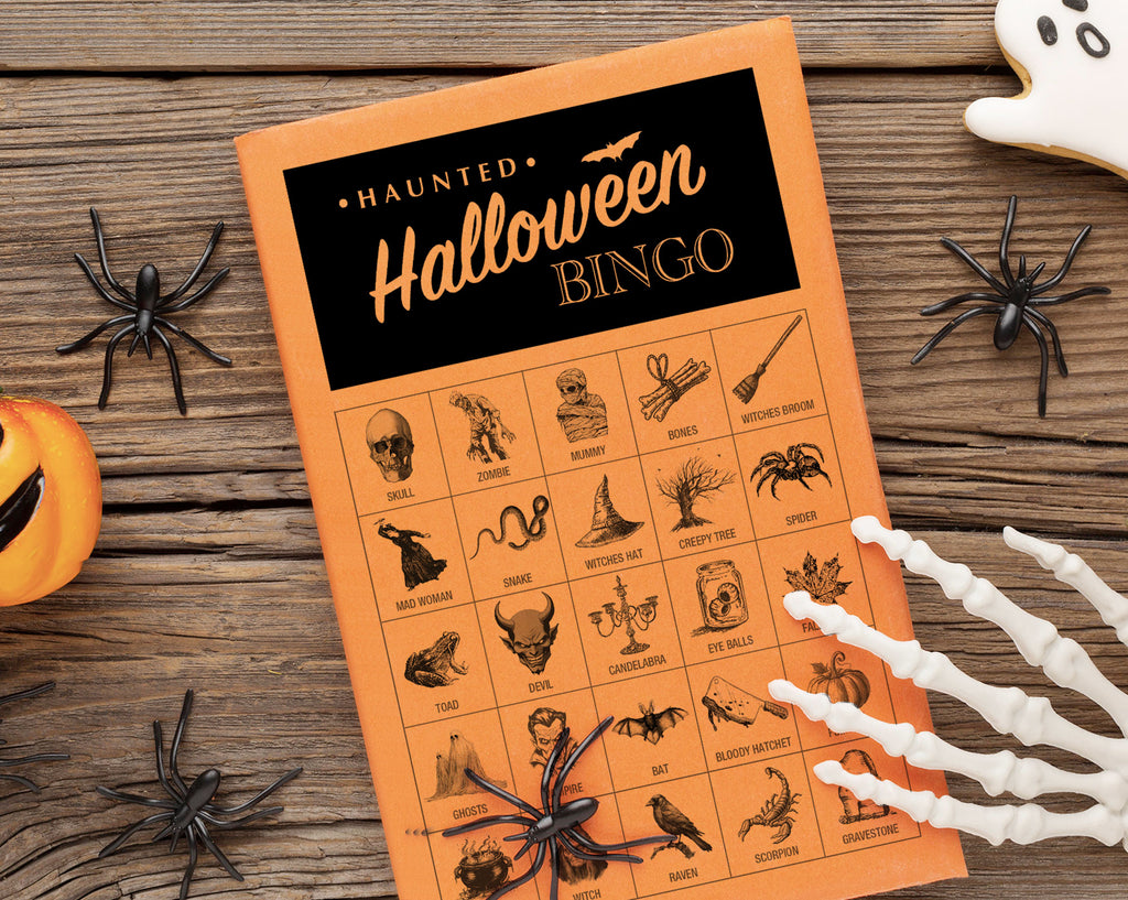 An orange and black 'Haunted Halloween Bingo' game card is displayed on a rustic wooden table surrounded by Halloween props including plastic spiders, a pumpkin, skeleton hands, and a ghost cookie.