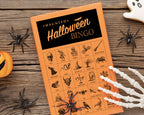 An orange and black 'Haunted Halloween Bingo' game card is displayed on a rustic wooden table surrounded by Halloween props including plastic spiders, a pumpkin, skeleton hands, and a ghost cookie.