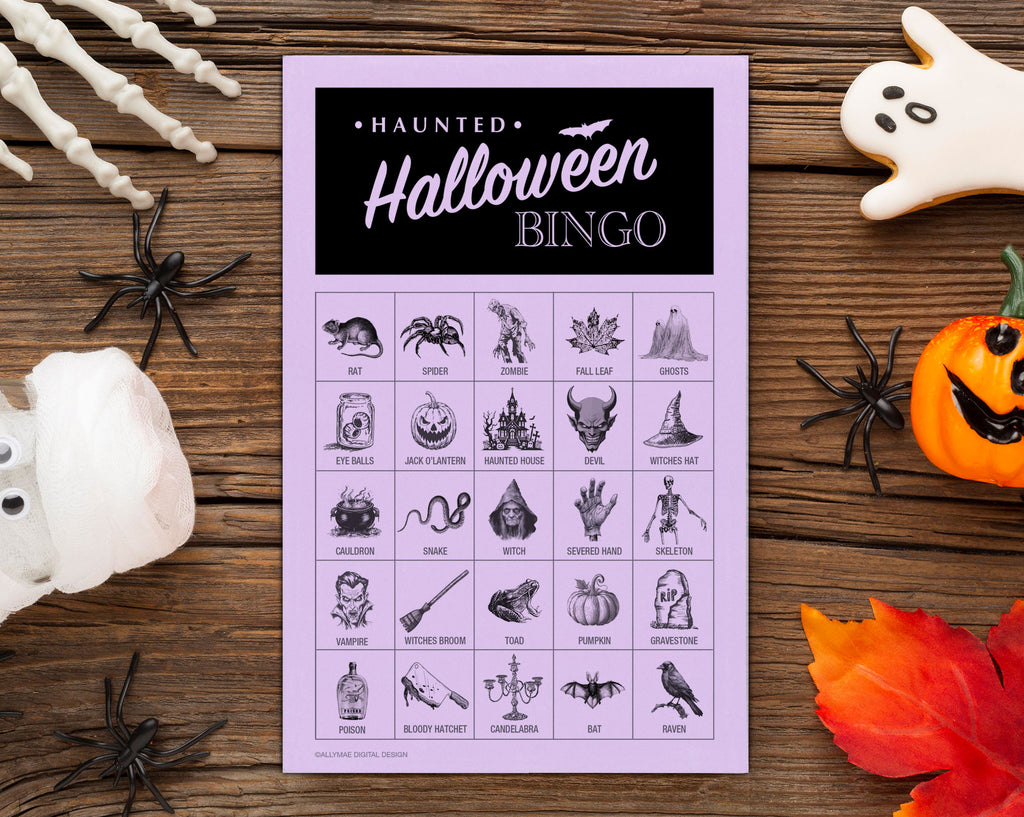 A Halloween-themed bingo card with a lavender background and black grid, surrounded by spooky props including plastic spiders, a jack-o'-lantern, a ghost cookie, skeleton bones, and autumn leaves on a wooden surface.