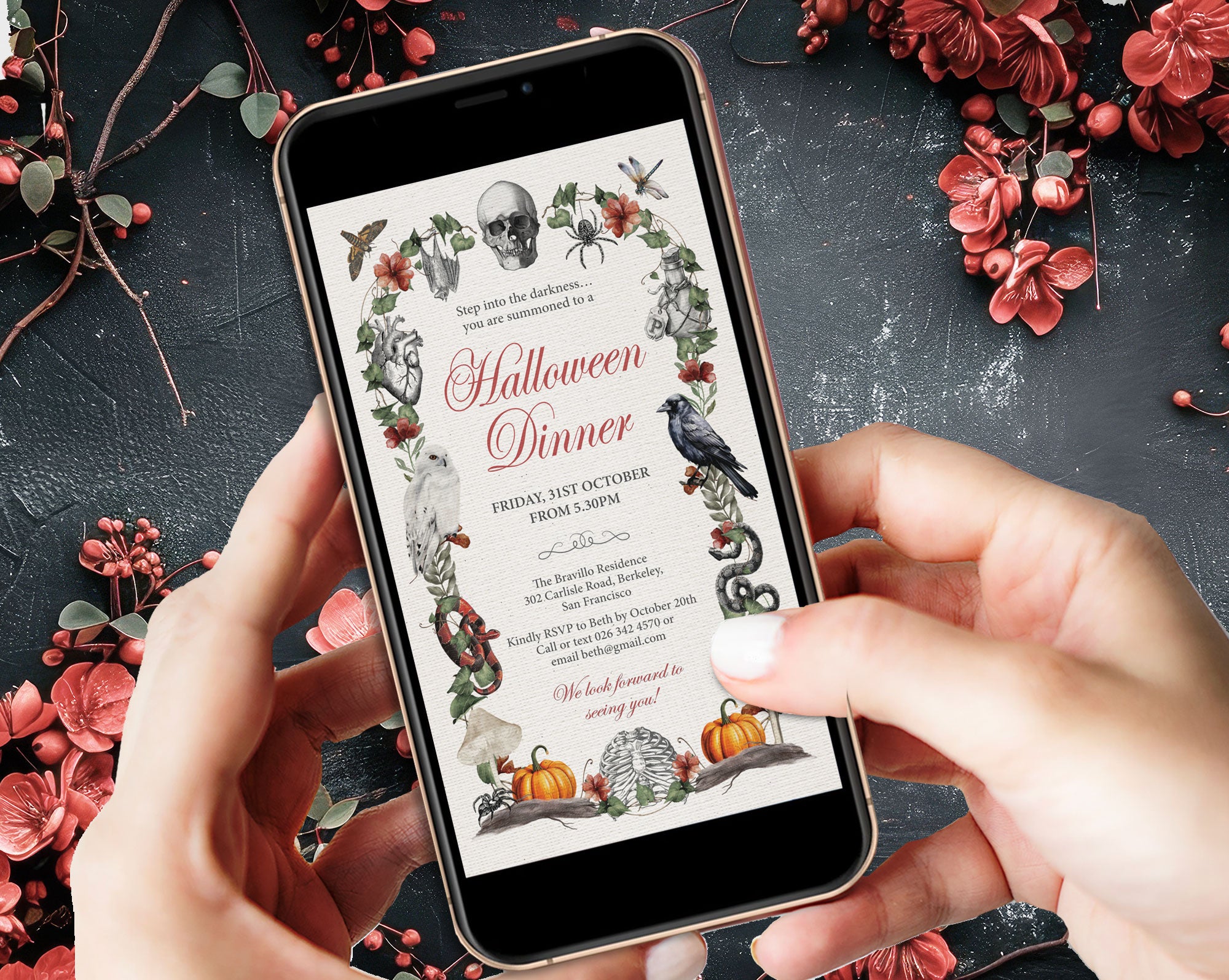 2 hands holding a smartphone displaying a Halloween dinner invitation card with gothic illustrations, set against a dark textured surface decorated with red flowers and berries.