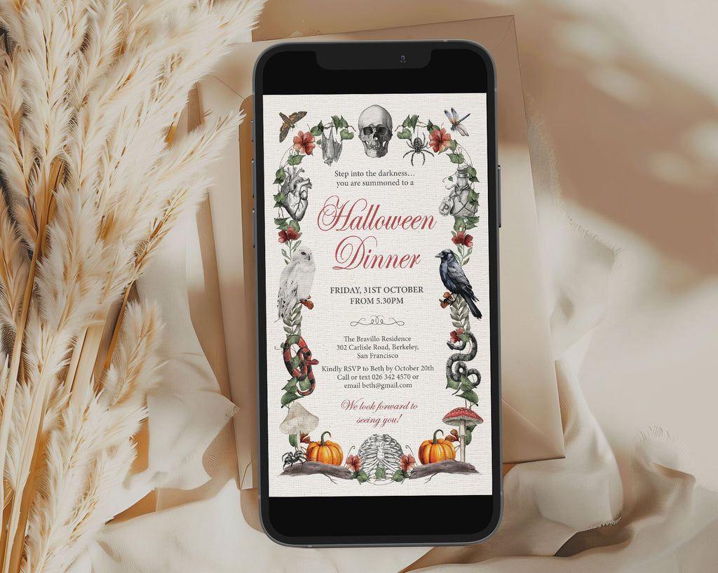 A smartphone displaying a Halloween dinner invitation design, styled on a beige surface with draped fabric and dried pampas grass. The invitation features a vintage-style illustration with a skull, bats, spiders, a raven, pumpkins, and floral accents.
