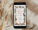 A smartphone displaying a Halloween dinner invitation design, styled on a beige surface with draped fabric and dried pampas grass. The invitation features a vintage-style illustration with a skull, bats, spiders, a raven, pumpkins, and floral accents.
