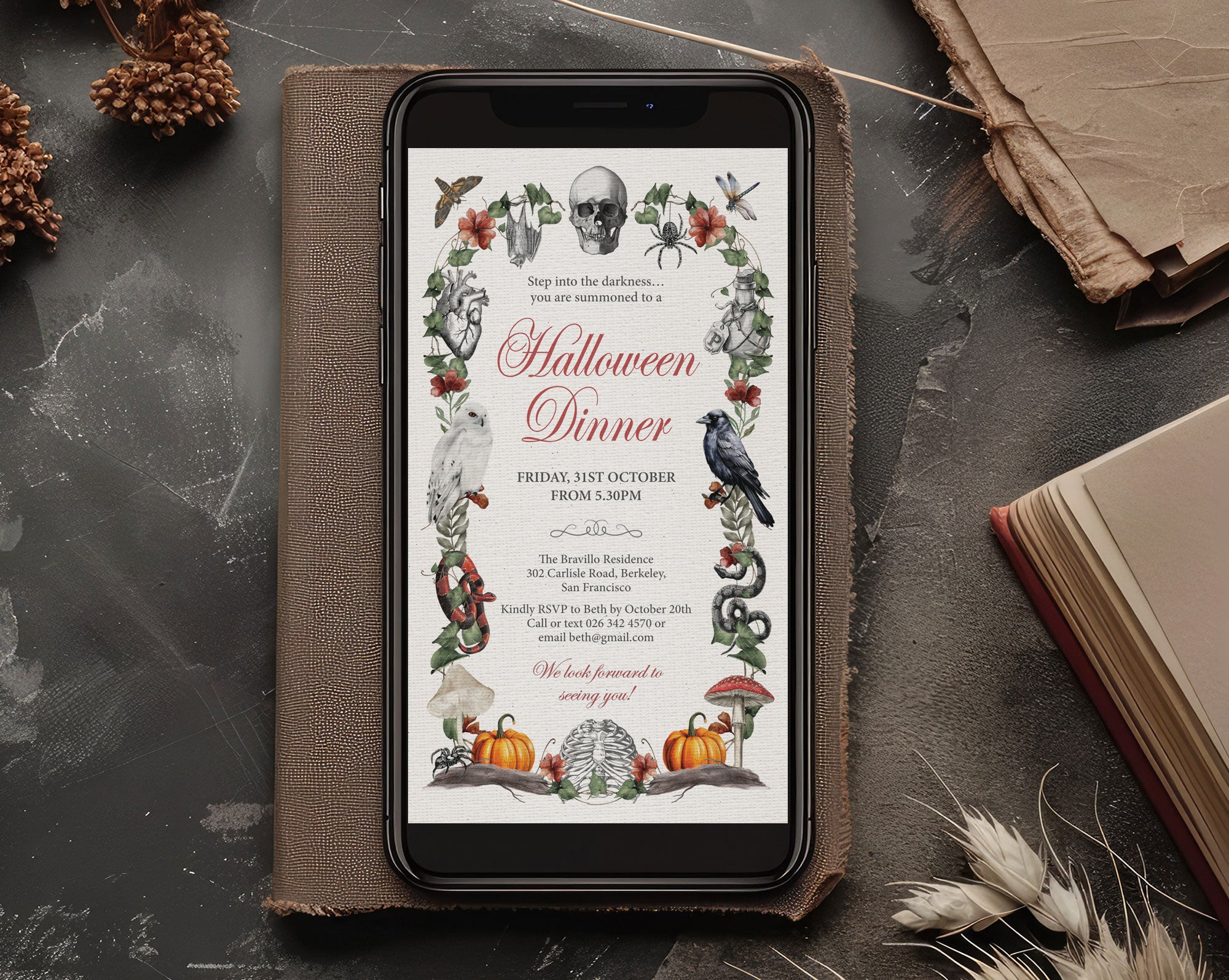 A smartphone displaying a Halloween dinner invitation card, placed on top of a brown leather journal. The scene is styled with dried wheat stalks, pinecones, and an old book on a dark textured tabletop.