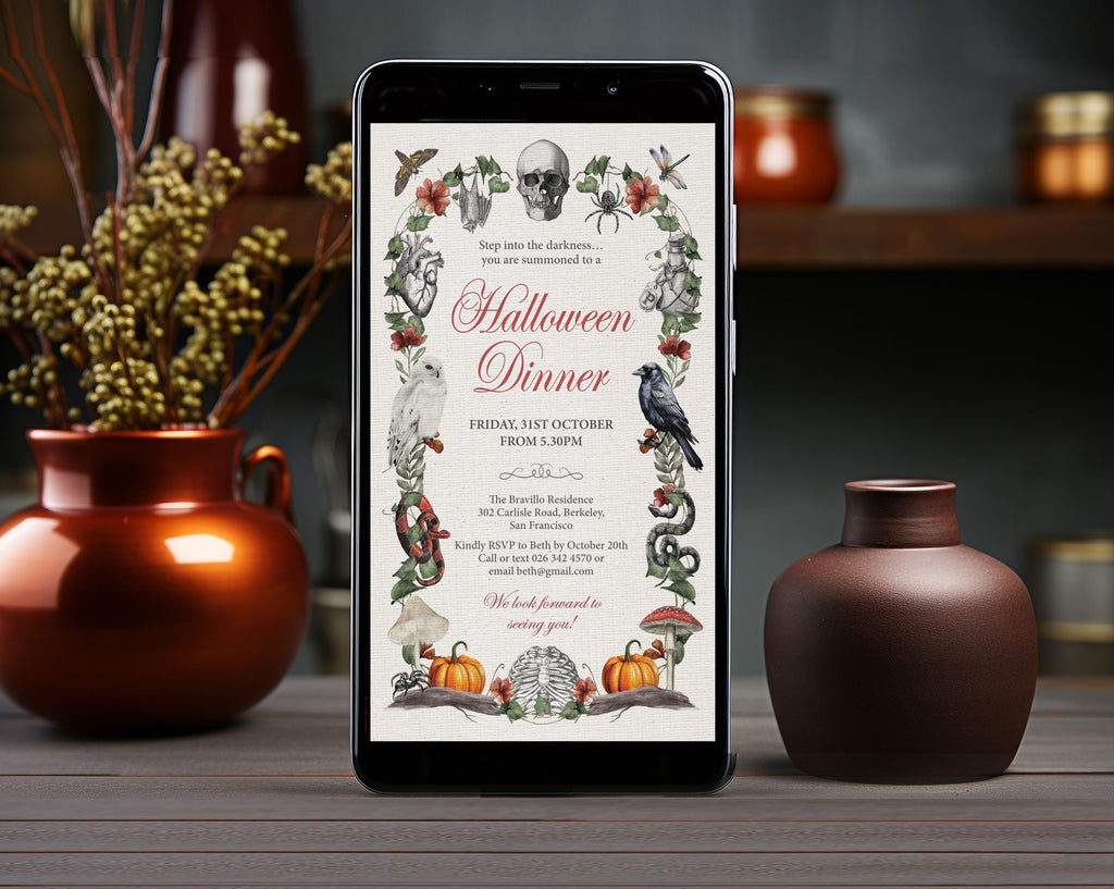 A smartphone centered on a wooden table, displaying a Halloween dinner invitation with vintage-style illustrations including a skull, crows, mushrooms, pumpkins, and floral borders. Bronze and brown ceramic vases and dried branches frame the scene in a styled indoor setting.