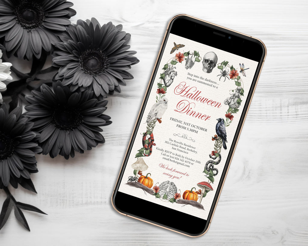 A smartphone lying on a white wood surface, displaying a Halloween dinner invitation design with illustrated skulls, pumpkins, crows, spiders, and floral borders. Black artificial flowers are arranged around the phone.