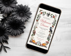 A smartphone lying on a white wood surface, displaying a Halloween dinner invitation design with illustrated skulls, pumpkins, crows, spiders, and floral borders. Black artificial flowers are arranged around the phone.