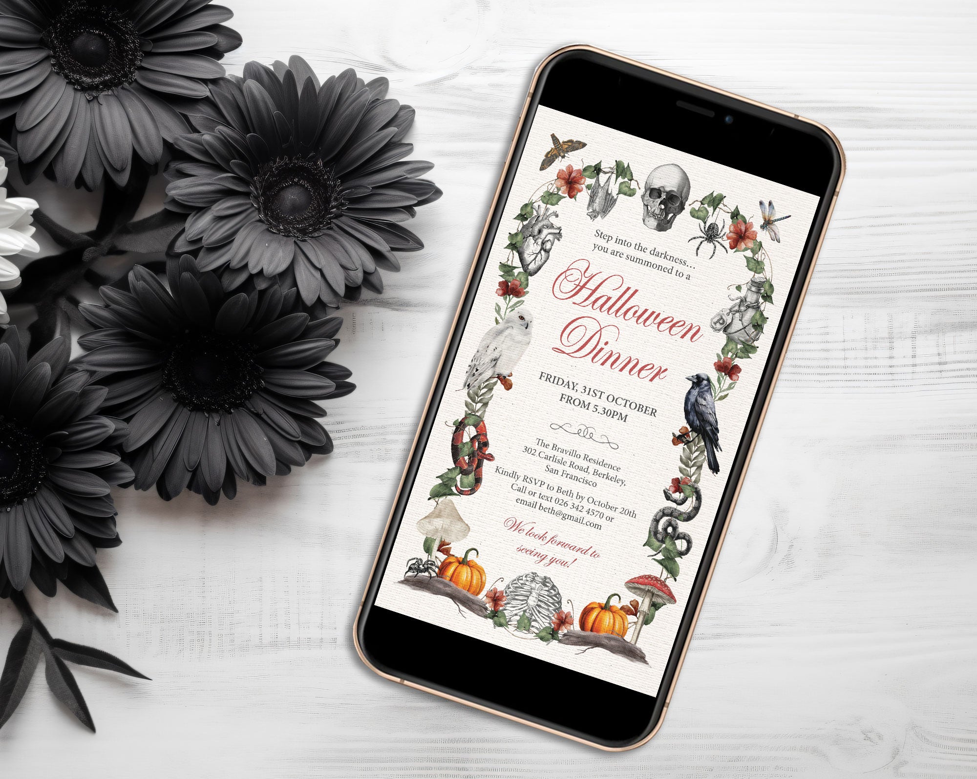 A smartphone lying on a white wood surface, displaying a Halloween dinner invitation design with illustrated skulls, pumpkins, crows, spiders, and floral borders. Black artificial flowers are arranged around the phone.