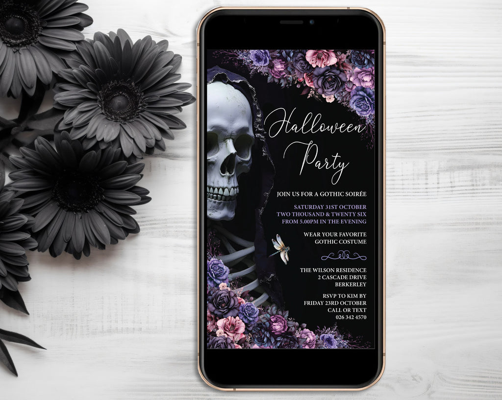 A smartphone displaying a gothic Halloween party invitation with a skull, flowers, and elegant text, placed on a white wood surface next to black daisy flowers.