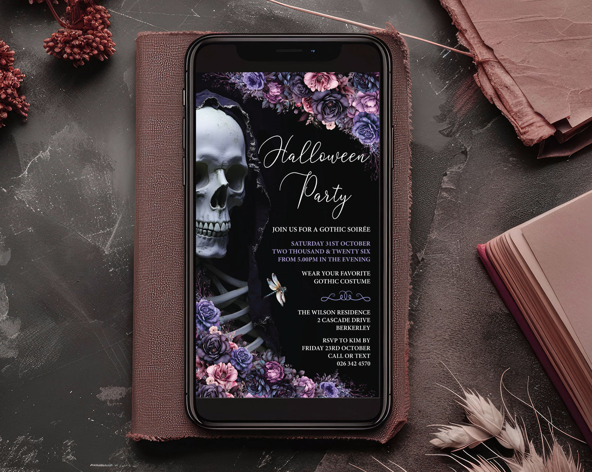 A smartphone lying on a brown leather folio, displaying a Halloween party invitation design with a skull, flowers, and gothic text. The scene is styled on a dark textured surface with dried flowers, a book, and a leather accessory nearby.