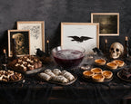 A Halloween-themed tabletop arrangement with a bowl of dark blood, mummy wraps, pumpkin pie tarts, bone-shaped cookies, candles, a skull, blackbirds, and framed gothic artwork against a dark textured wall.