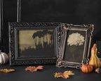 Two ornate picture frames displayed on a dark surface against a black background. The larger left frame is black with carved detailing and contains a dark landscape print.