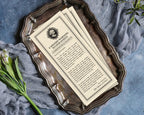 Vintage-style printed Halloween victorian society page with ornate black text and border design, placed on a distressed silver tray with draped blue-gray fabric and green stems around it.