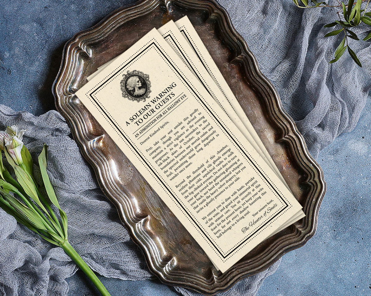 Vintage-style printed Halloween victorian society page with ornate black text and border design, placed on a distressed silver tray with draped blue-gray fabric and green stems around it.