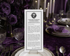 A vertical printed halloween card reading 'A Solemn Warning to Our Guests' is displayed upright on a table setting with purple flowers, plates, candles, and silverware in the background.