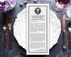 A printed Halloween Victorian style card with creepy skull cameo reading 'A SOLEMN WARNING TO OUR GUESTS' is laid flat on a dark marble surface, surrounded by white plates, silverware, a glass of red wine, and purple flowers.