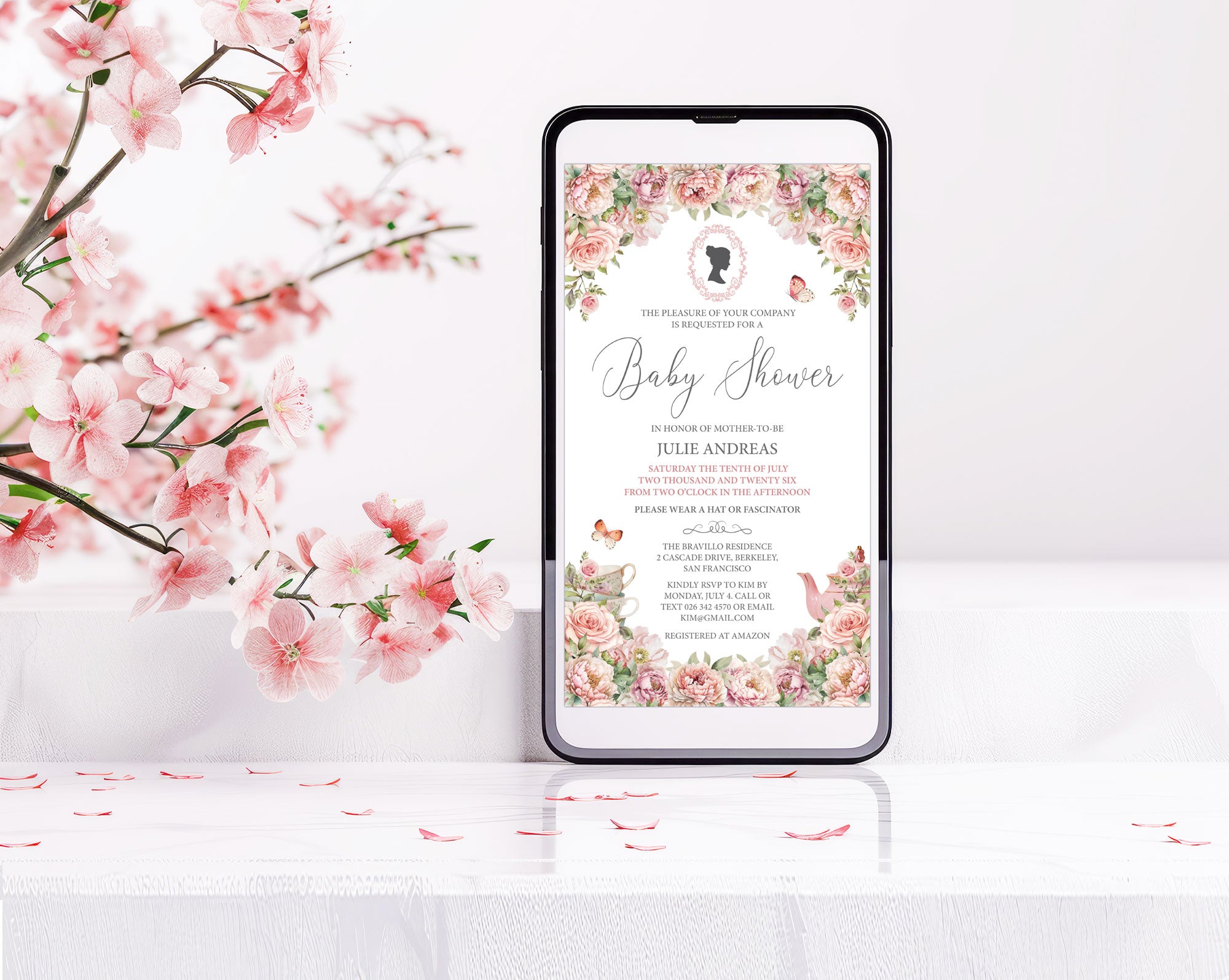 a black mobile phone with a pink flower baby shower tea invitation showing on the screen, leaning up against a white wall with some pink blossoms