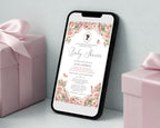 a black mobile phone showing a pink flower baby shower regency style invitation leaning up against a white wall next to some pink wrapped gifts
