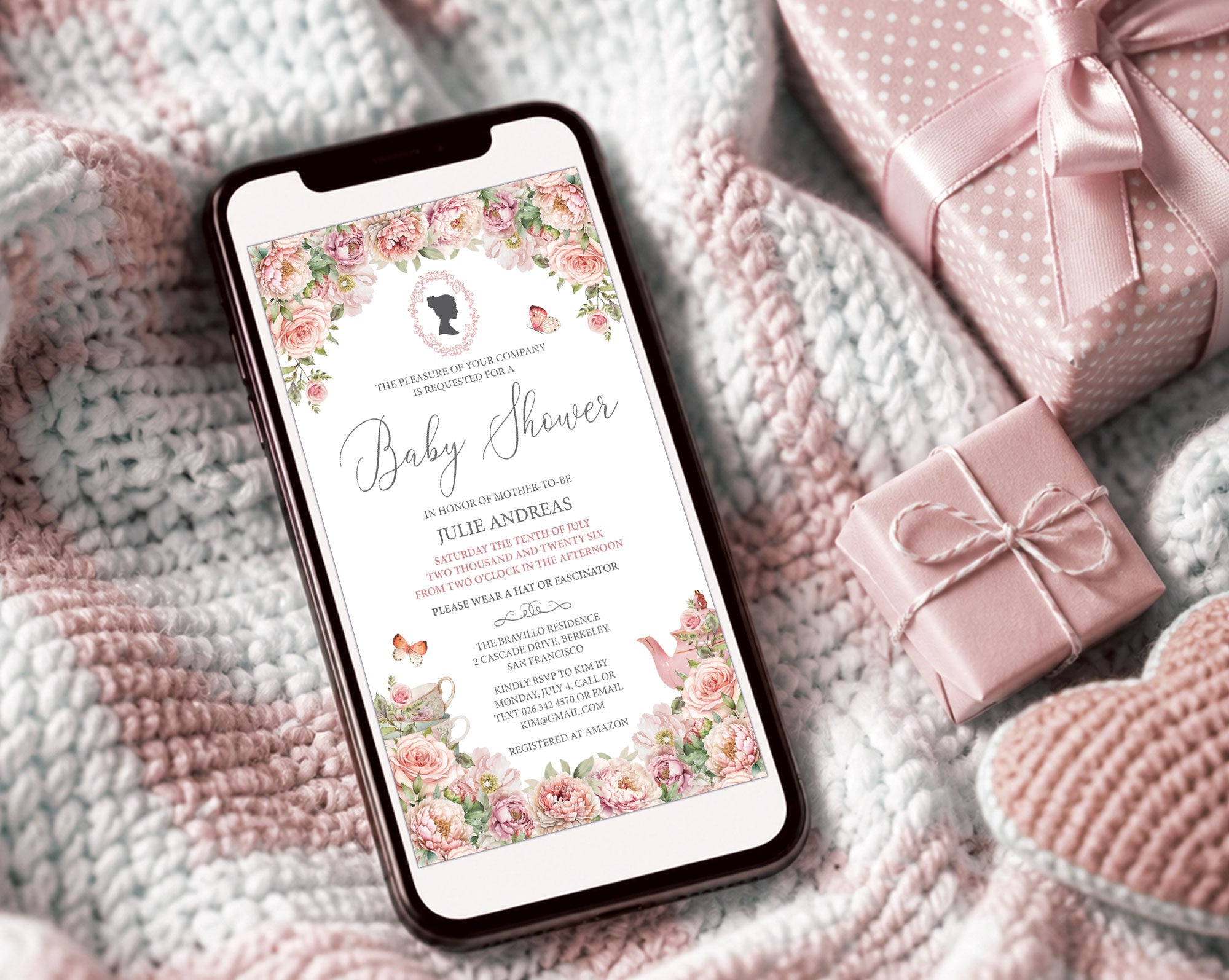 pretty pink regency style baby shower invitation showing on the screen of a mobile phone, laying on a baby blanket next to some pink wrapped gifts