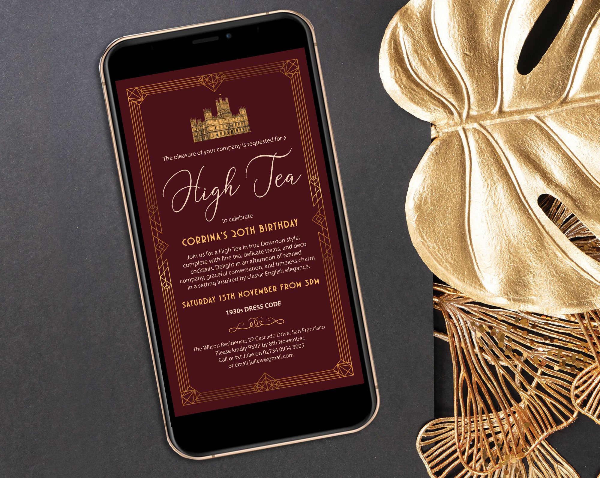 black mobile phone with an art deco style burgundy and gold high tea invitation laying on a grey background next to gold leaf and decor